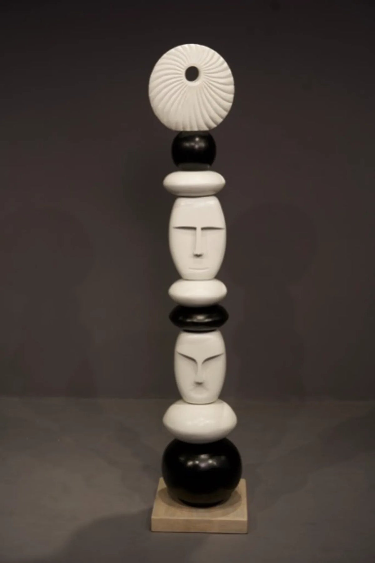 A modern abstract sculpture featuring stacked organic shapes and stylized faces in black and white, standing on a light beige square base against a dark gray studio background.
