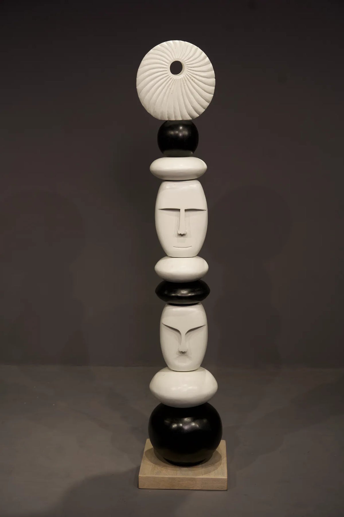 A modern abstract sculpture made of stacked organic shapes and stylized faces in glossy black and white, standing on a small beige square base against a dark gray studio background.