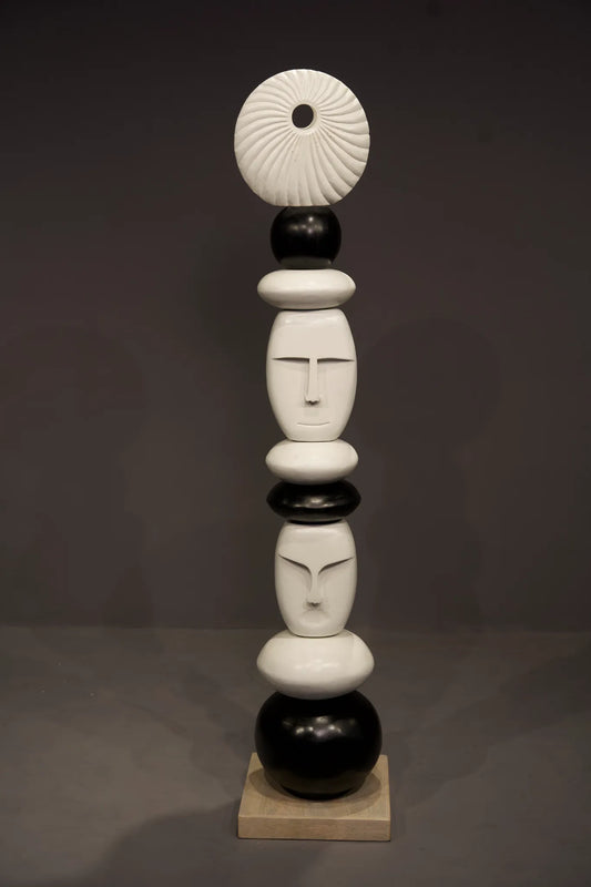 A modern abstract sculpture made of stacked organic shapes and stylized faces in glossy black and white, standing on a small beige square base against a dark gray studio background.
