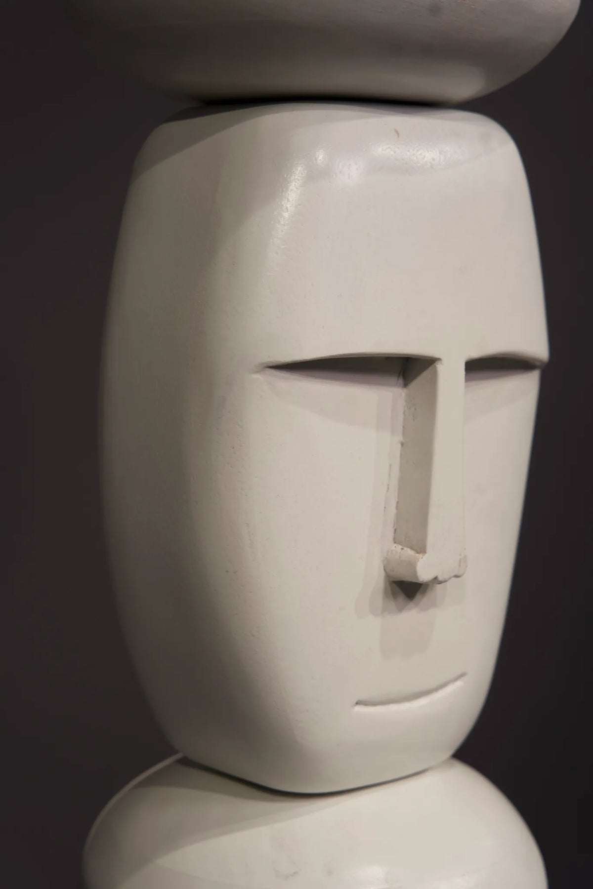 A close-up of a white sculptural face sculpture with a minimalist, stylized expression, set against a dark gray background.