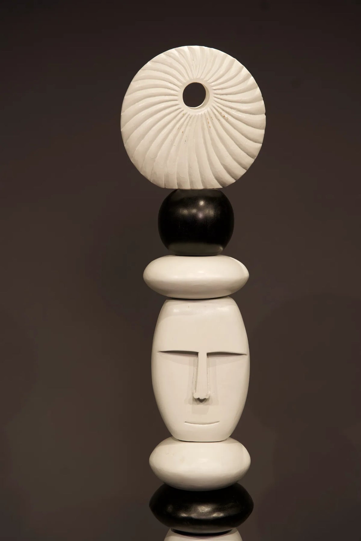 A modern abstract sculpture made of stacked organic shapes, featuring a white ribbed circular top, a black sphere, a white oval, a white face-like form with a simple face, and two black bases, set against a dark gray background.