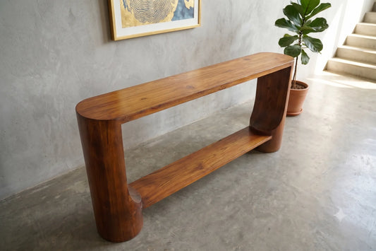 A mid-century modern wooden console table with a rich warm brown finish, set in a minimalist interior with a concrete floor, textured gray wall, and a potted plant.