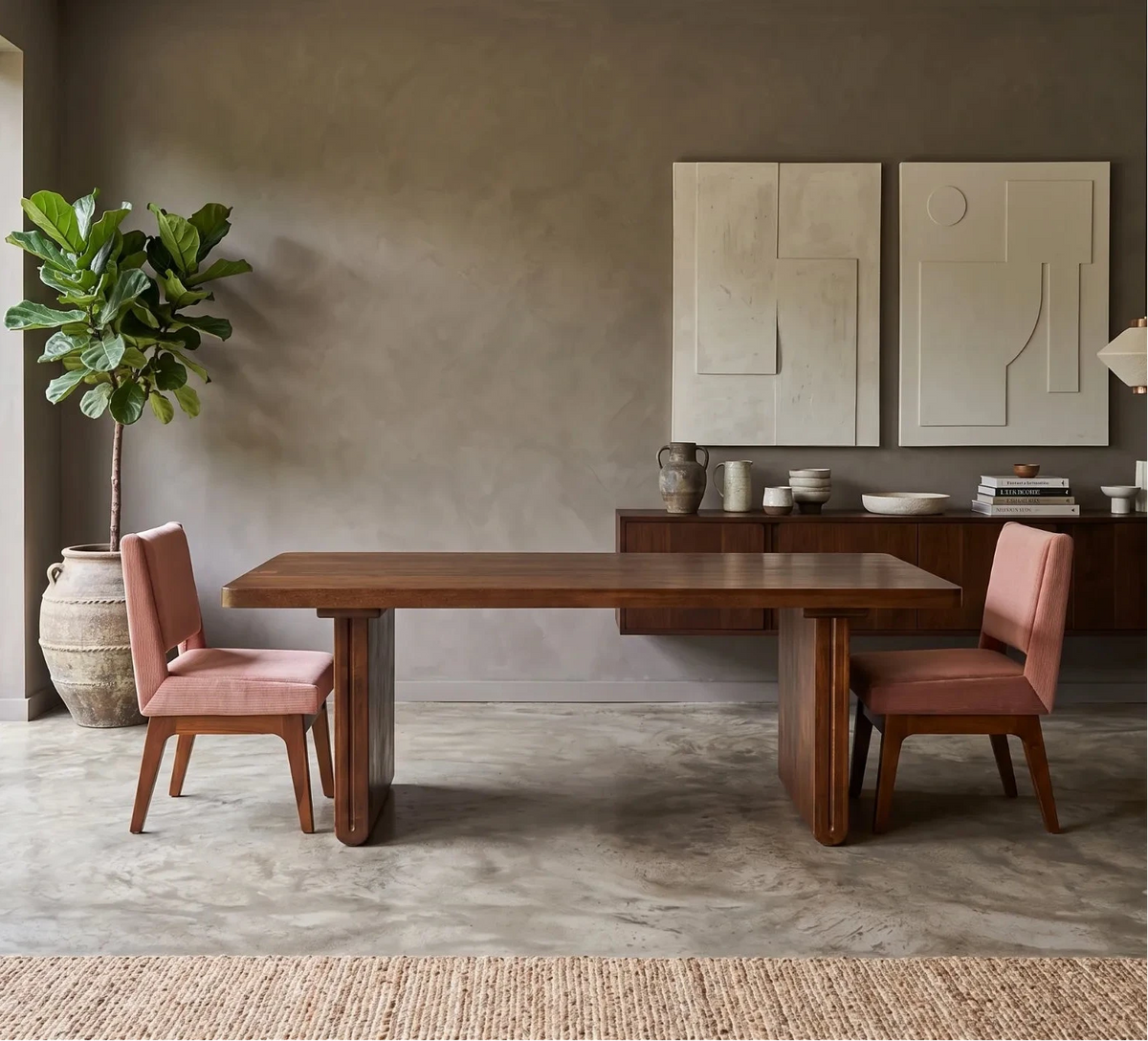A modern dining room setup featuring a rectangular dark wood table with two pink upholstered chairs, set in a minimalist room with a concrete floor, textured gray walls, and a large potted plant by a window.