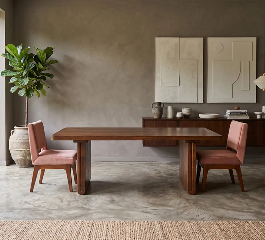 A modern dining room setup featuring a rectangular dark wood table with two pink upholstered chairs, set in a minimalist room with a concrete floor, textured gray walls, and a large potted plant by a window.