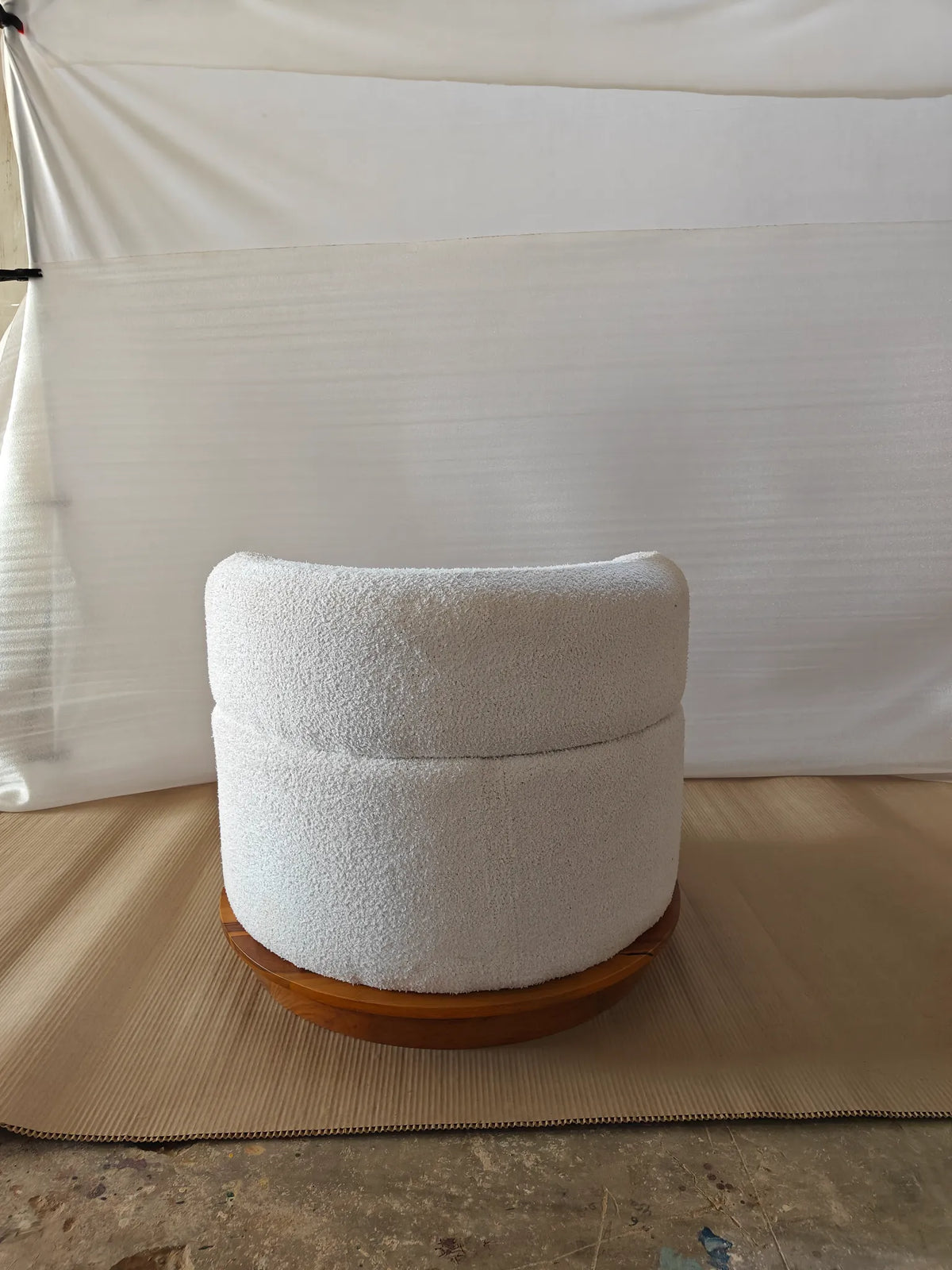 A modern cylindrical pouf or ottoman with a textured off-white bouclé fabric cushion on a round wooden base, set against a white fabric backdrop.