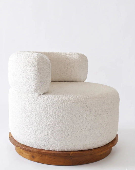 A modern low-profile barrel chair upholstered in textured off-white bouclé fabric, featuring a rounded backrest and a circular natural wood base, photographed against a seamless white background.