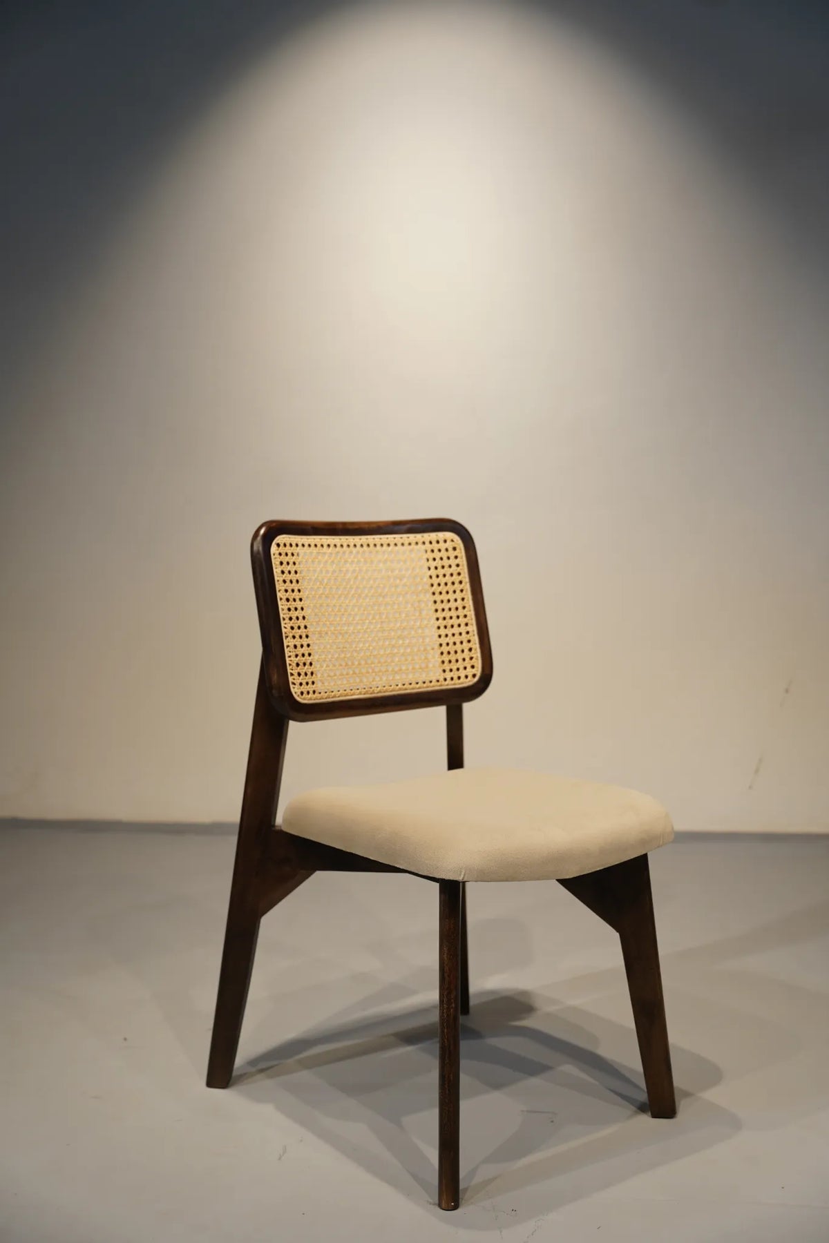 A mid-century modern dining chair with a dark wood frame, a woven cane backrest, and a cream upholstered seat cushion, set against a neutral gray studio background with a spotlight effect.