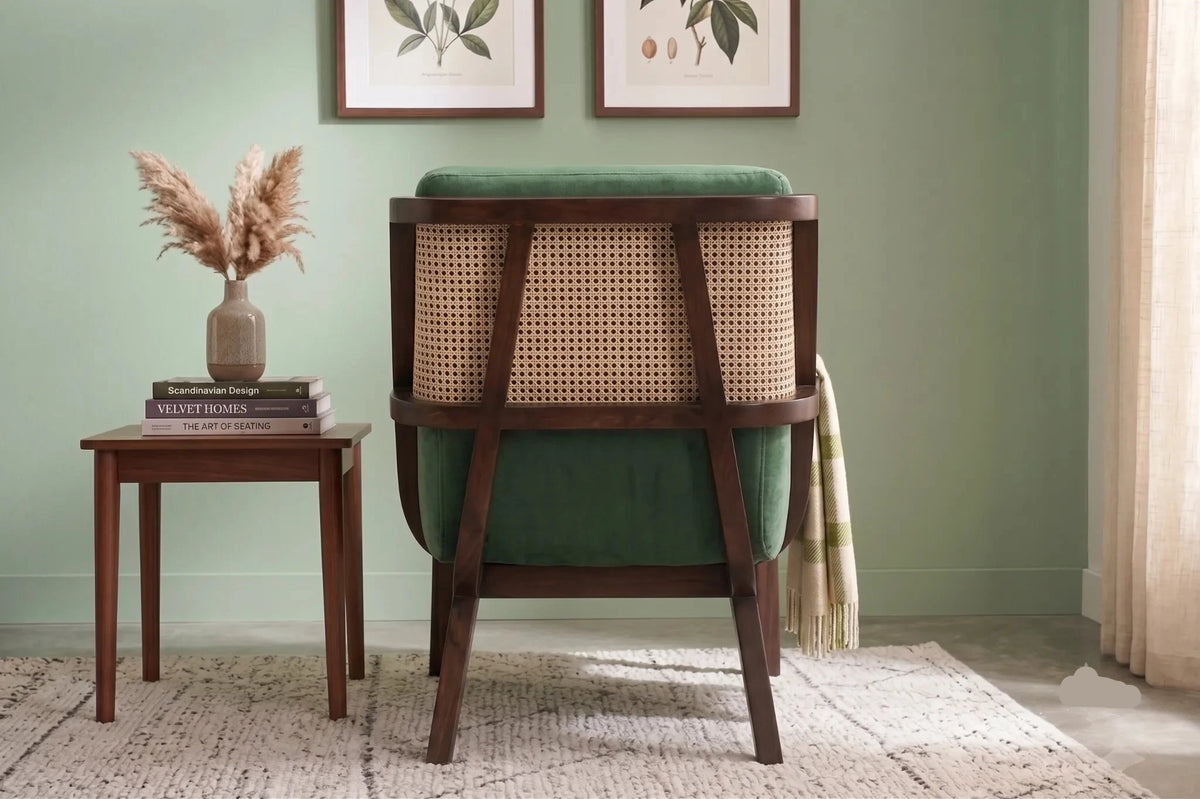 A wooden armchair with a green velvet seat and back, featuring a woven cane backrest, shown in a styled room setting next to a matching wooden side table.