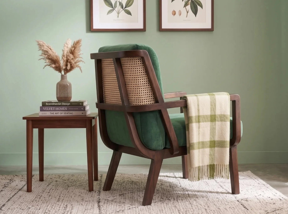 A mid-century modern accent chair with dark brown wood, a green velvet seat and back cushion, and a woven cane backrest, styled in a room with a sage green wall, a side table, and a cream plaid throw blanket.