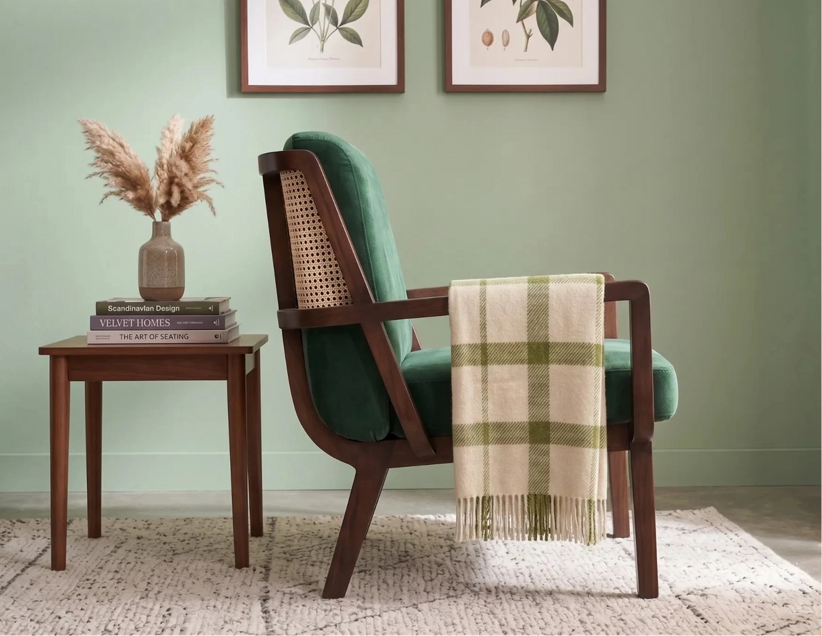 A mid-century modern accent chair with deep green velvet upholstery and a woven cane backrest, styled in a room with a sage green wall, a small wooden side table, and a cream plaid throw blanket draped over the arm.