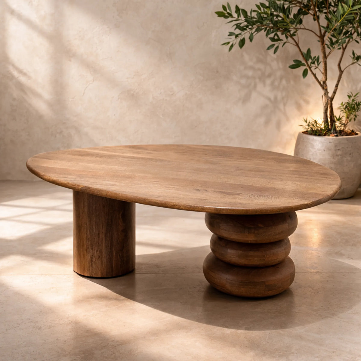 An organic-shaped wooden coffee table with a thick round top and a stacked, chunky pedestal base, set in a warm, sunlit room with a textured beige wall and a potted plant in the background.