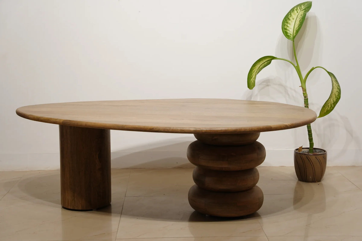 An oval wooden coffee table with a light natural wood top and a dark brown sculptural stacked pedestal base, shown in a minimalist room setting next to a potted plant.
