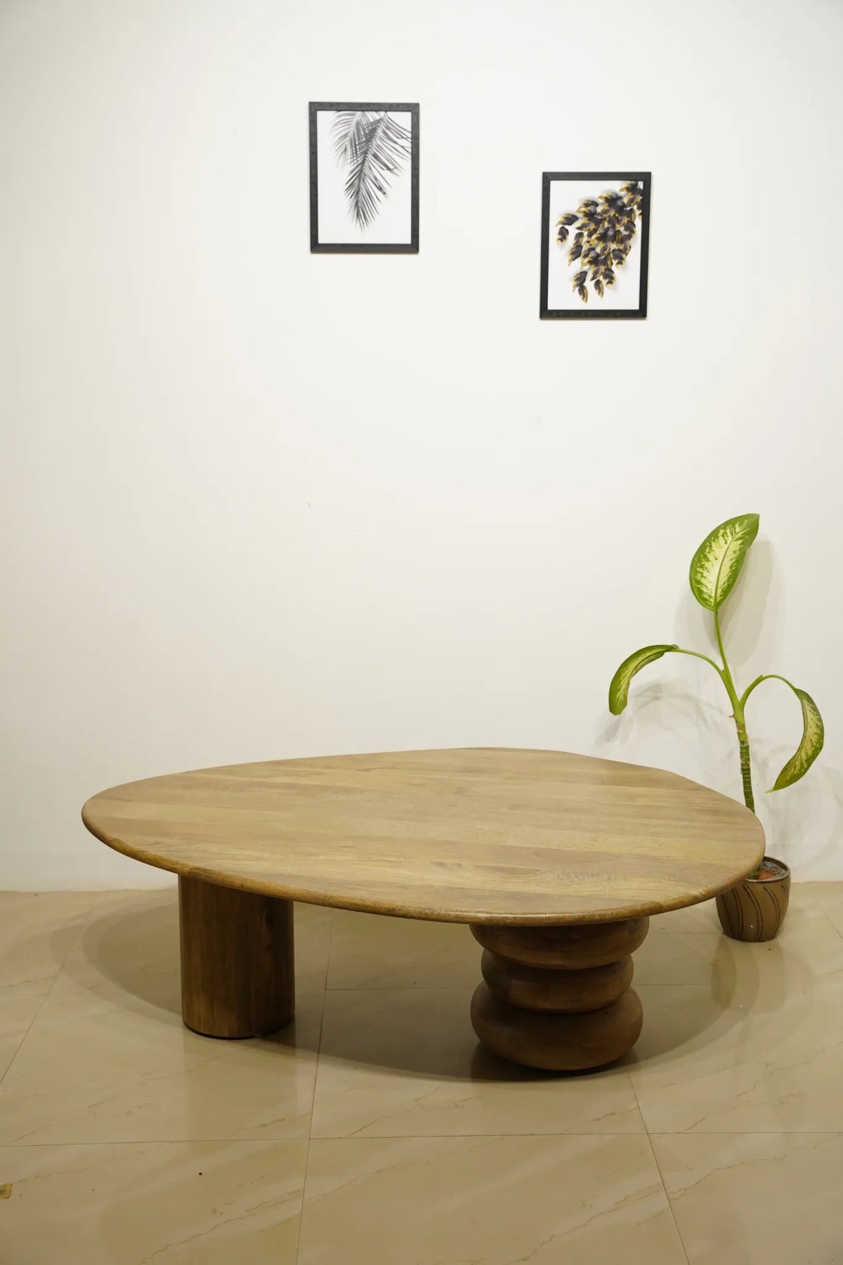 A low, organic-shaped wooden coffee table with chunky cylindrical legs stands in a minimalist room with a white wall, two framed botanical prints, and a potted green plant.