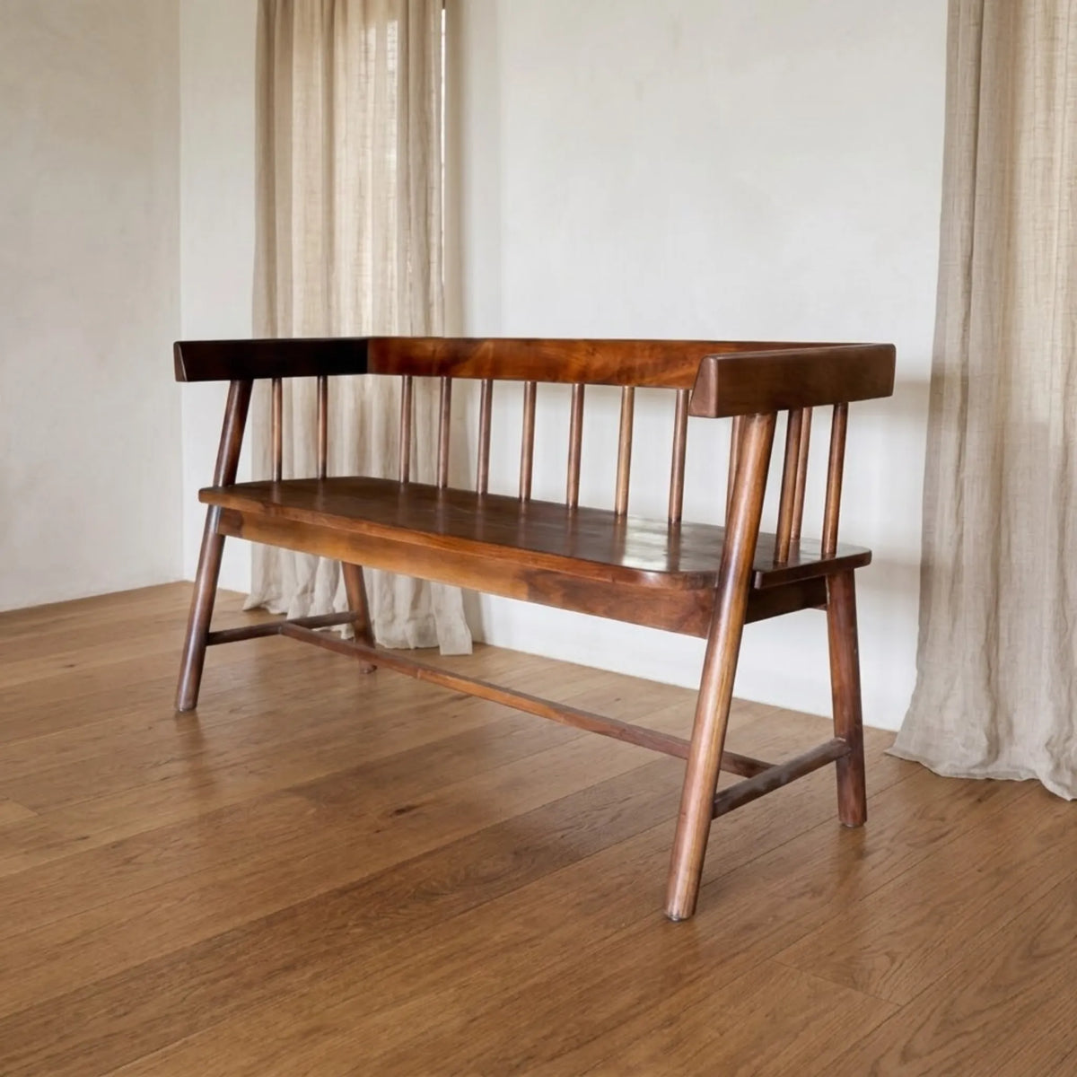 A mid-century modern wooden bench with a spindle backrest, shown in a warm brown finish on a wooden floor in front of a white wall with neutral curtains.