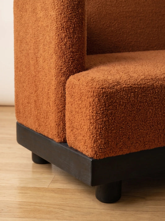 Close-up of a textured terracotta orange bouclé upholstered sofa corner with a dark wood base and cylindrical legs on a light wood floor.