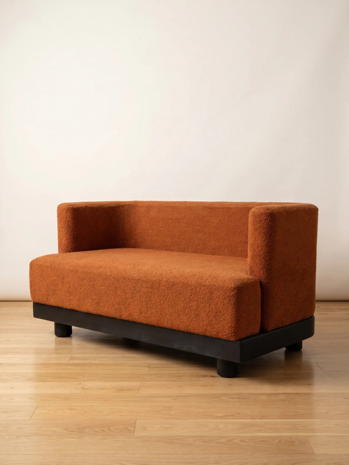 A modern two-seater sofa with a textured rust-orange bouclé fabric and a dark wood base, set against a plain off-white wall and light wood floor.