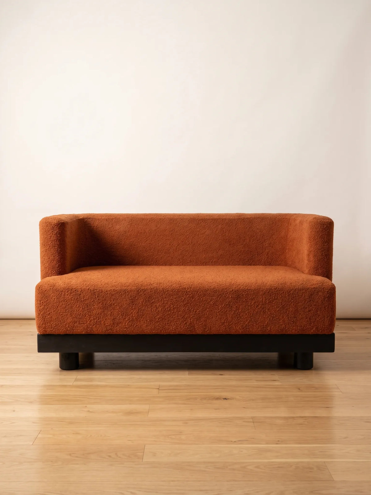 A modern two-seater sofa upholstered in a textured rust-orange bouclé fabric with a dark wood base, set against a plain off-white wall and light wood floor.